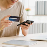 Woman using smartphone buying online shopping by credit card while wear sweater sitting on desk in living room at home